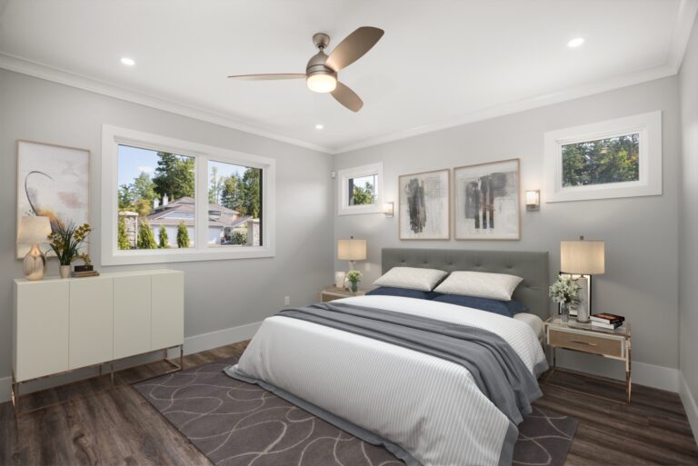 Modern bedroom interior with two neatly made beds, gray tufted headboard, matching nightstands, and abstract wall art. Large windows show a sunny suburban neighborhood. Ceiling fan above and recessed lights illuminate the room.