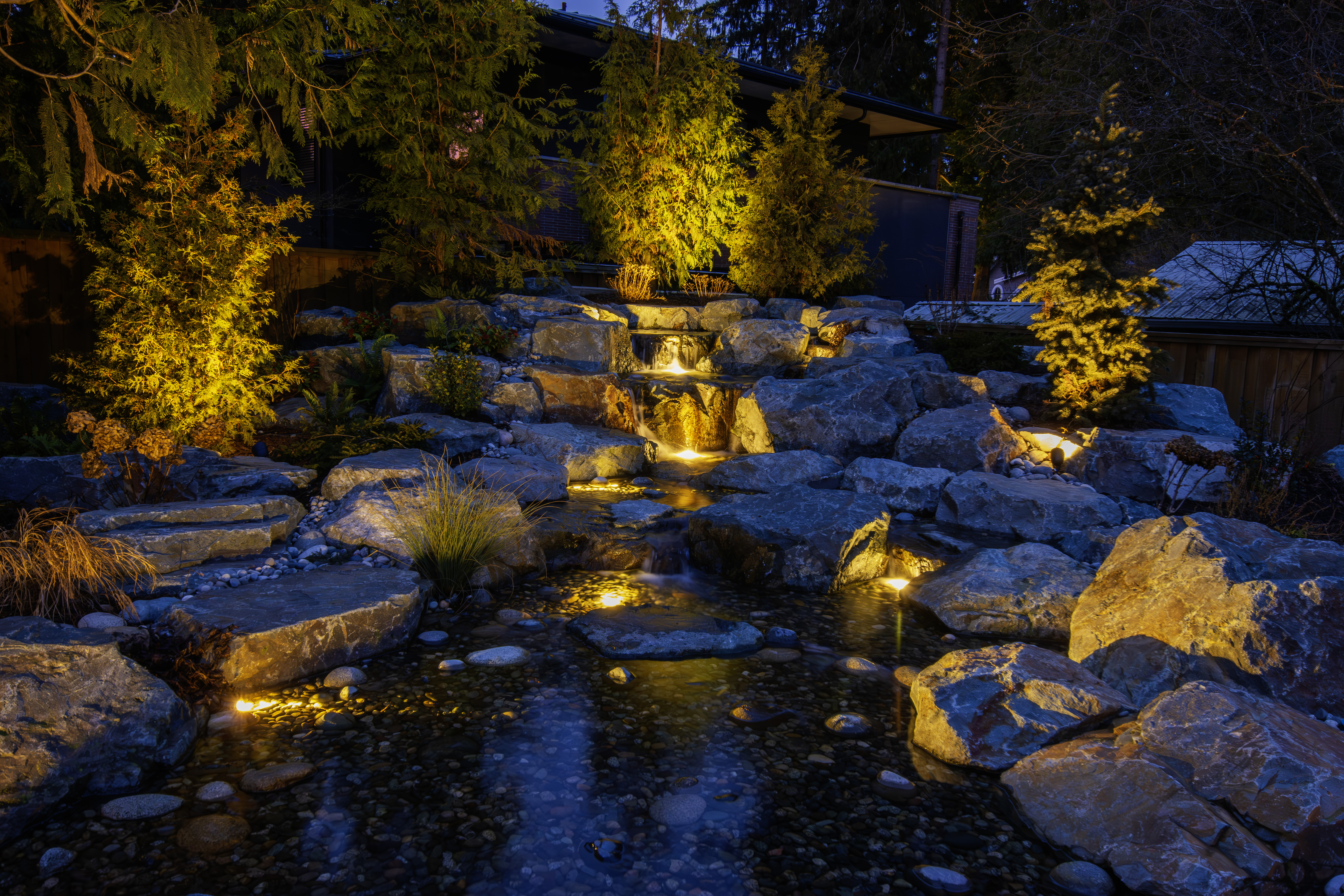 Night scene of a rock garden with cascading lights and a small waterfall beside a house.