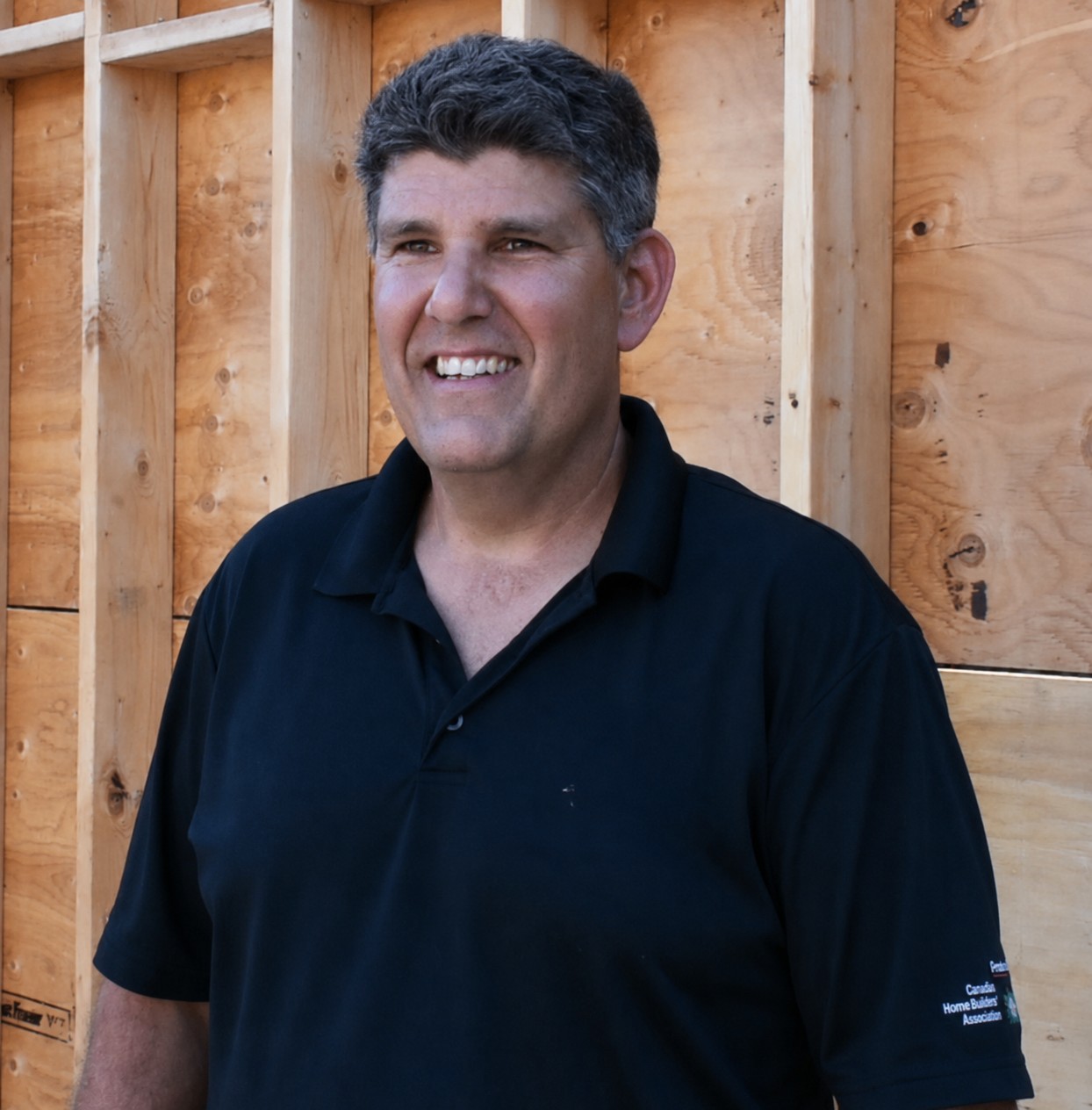Smiling man in a dark polo shirt stands in front of unfinished wooden framing at a construction site.
