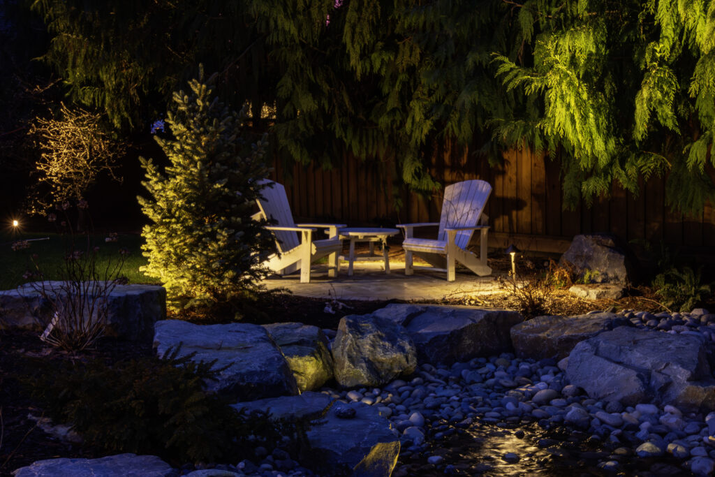 Night scene of a backyard seating area with two white Adirondack chairs around a small table, under dense overhanging branches lit by warm garden lights.