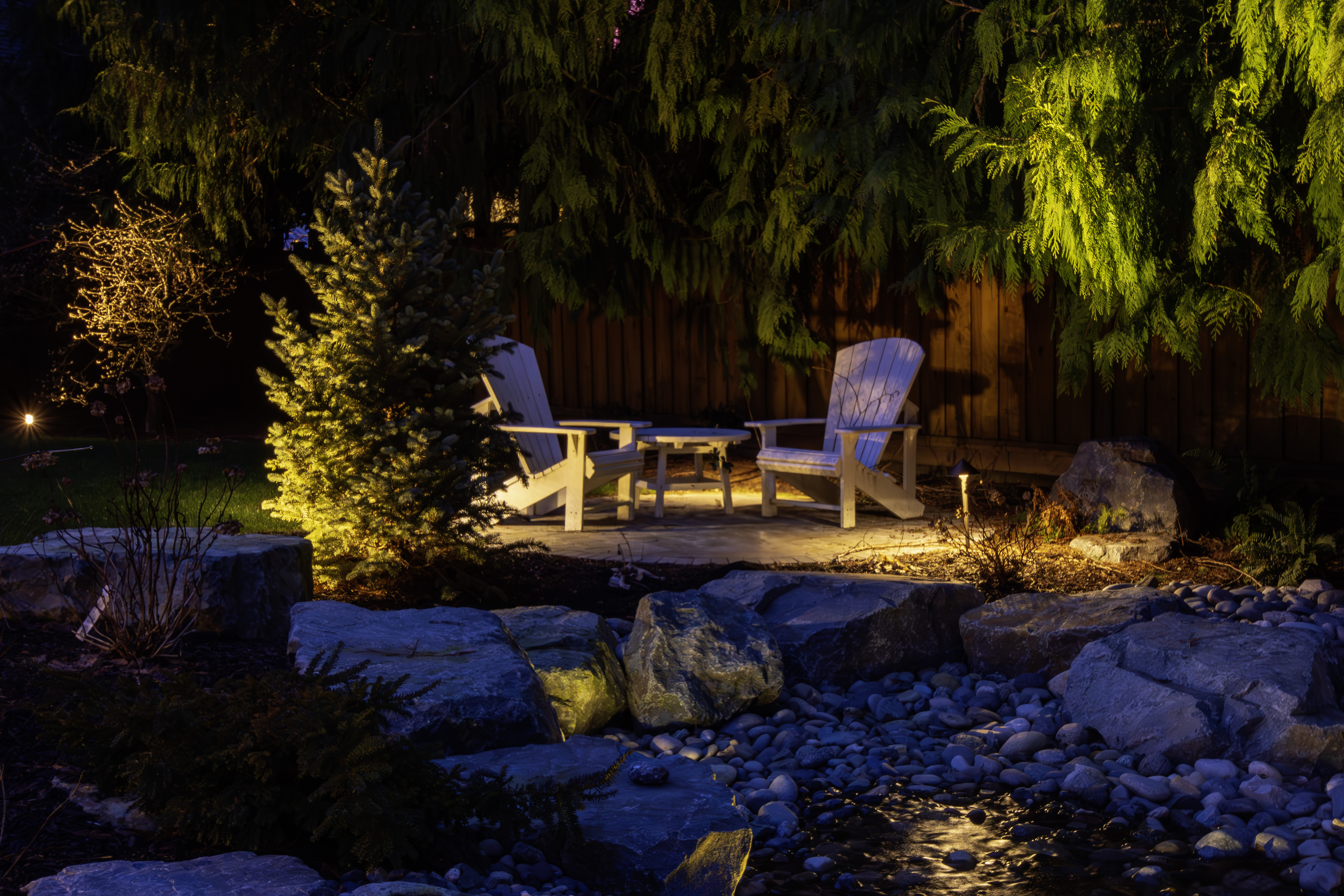 Night garden seating area with white wooden chairs and a small table beneath hanging evergreen branches, warm outdoor lighting murkily illuminating the scene.